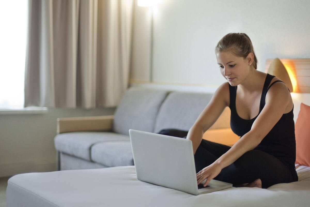 Eine Person benutzt einen Laptop im Bett in einem Zimmer mit Sofa und Lampe.