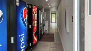 Un couloir doté de distributeurs automatiques, notamment de Pepsi et de Coca-Cola, et une porte donnant sur un espace lumineux.