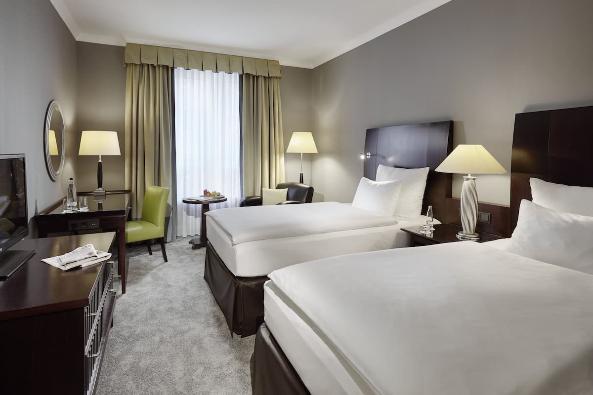 Ein Hotelzimmer mit zwei Betten, einem Schreibtisch, einem Stuhl, einem kleinen Tisch und einem Fenster mit Vorhängen.