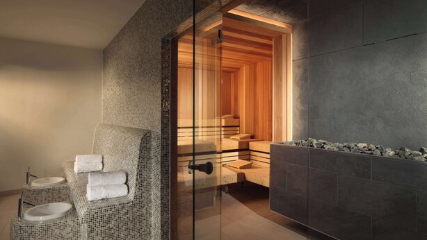 A modern bathroom with a glass-enclosed shower, a wicker sofa with towels, and a stone-tiled wall.