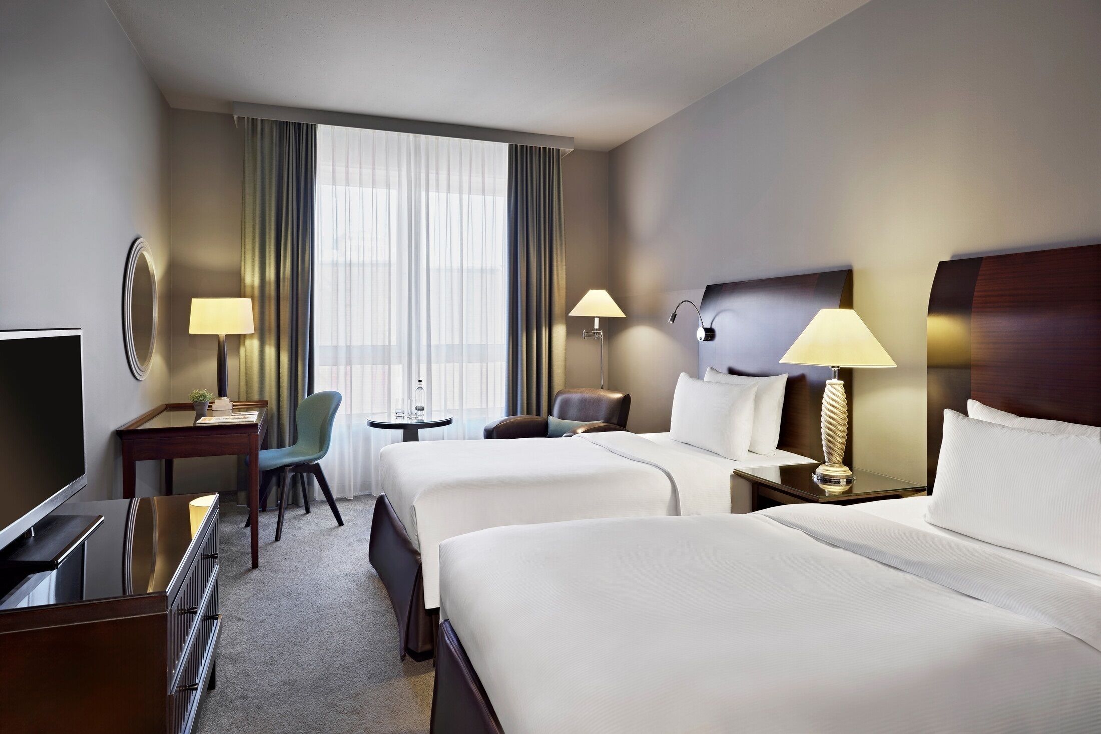 A hotel room with two beds, a desk, a chair, a TV, and a window with curtains.