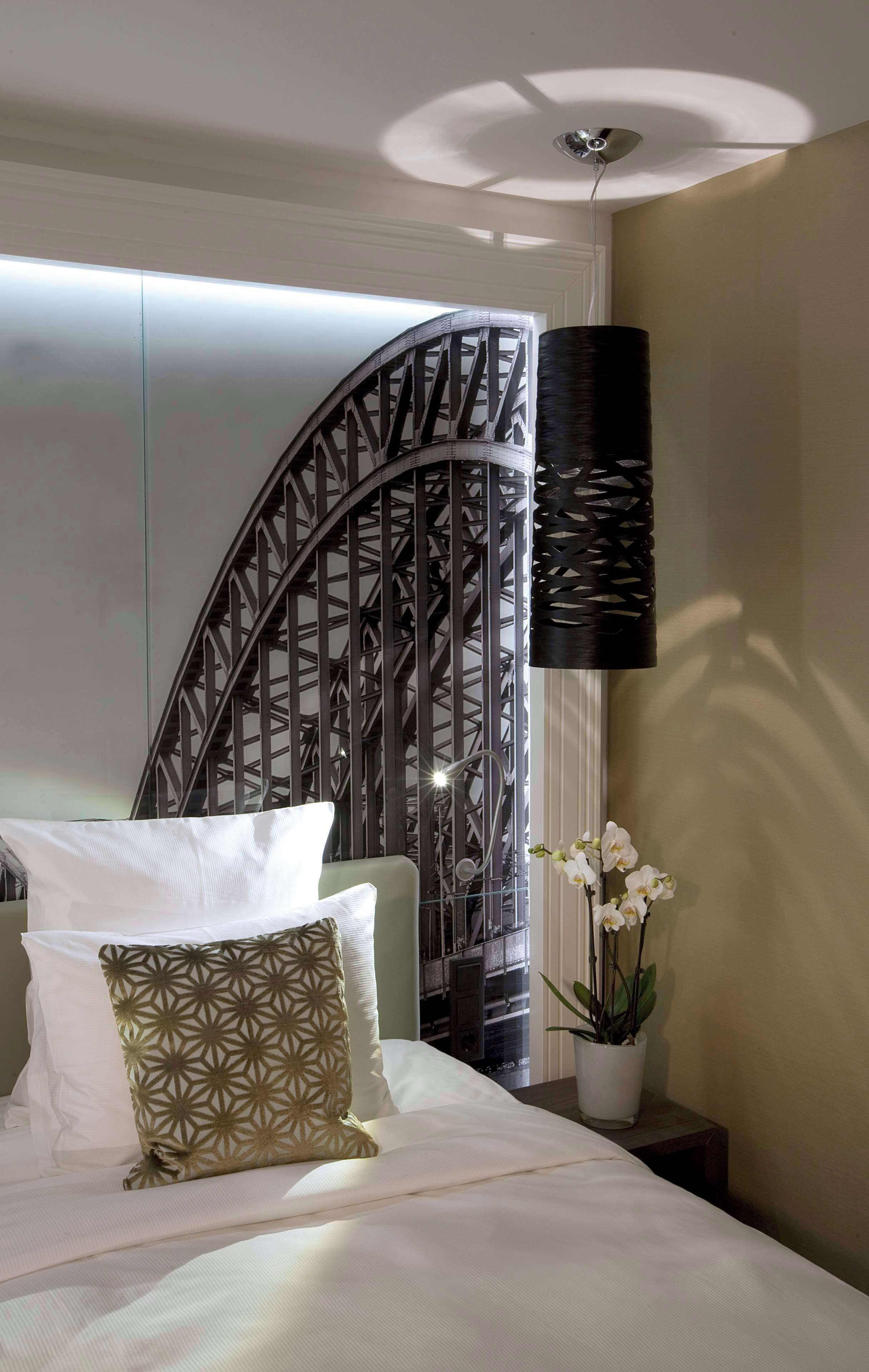 Ein ordentlich bezogenes Bett mit weißen und gemusterten Kissen, ein dekorativer Metallraumteiler, eine Hängelampe und eine Orchidee im Blumentopf.