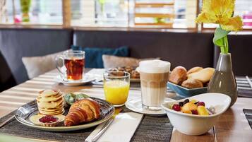 A breakfast setting with pancakes, croissants, a croissant with jam, a bowl of fruit, a glass of orange juice, a glass of milk, and a vase with flowers.