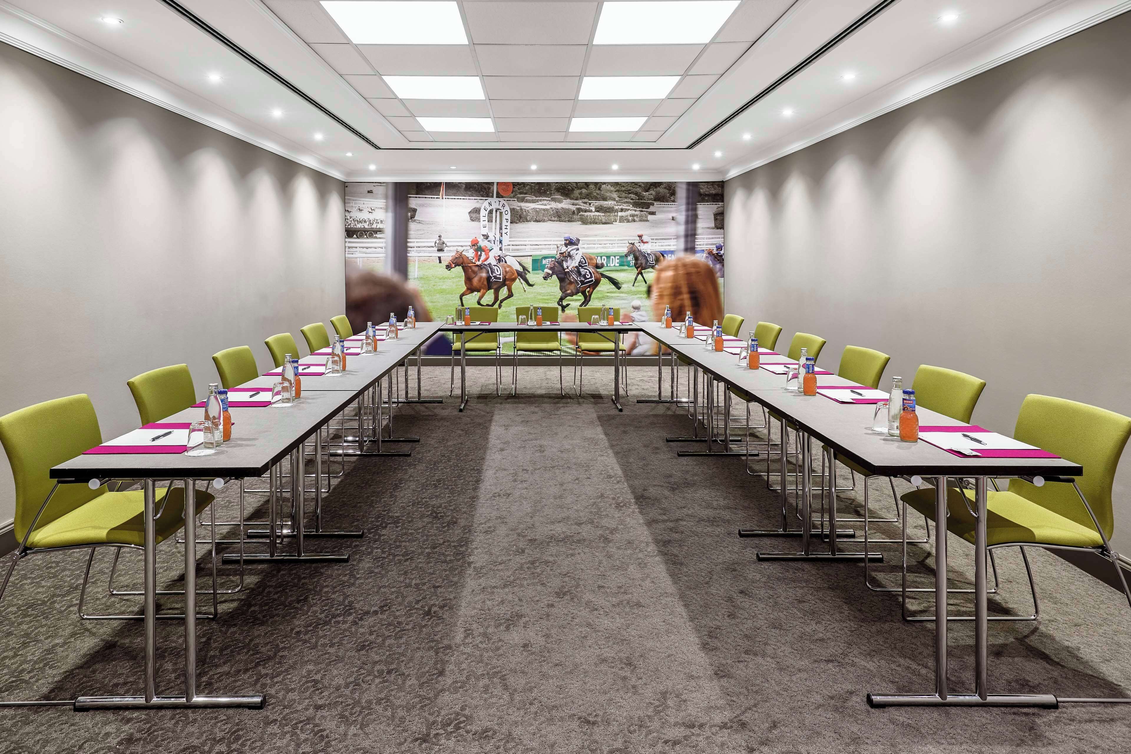 Een vergaderruimte met een lange tafel, groene stoelen en een vloerbedekking.