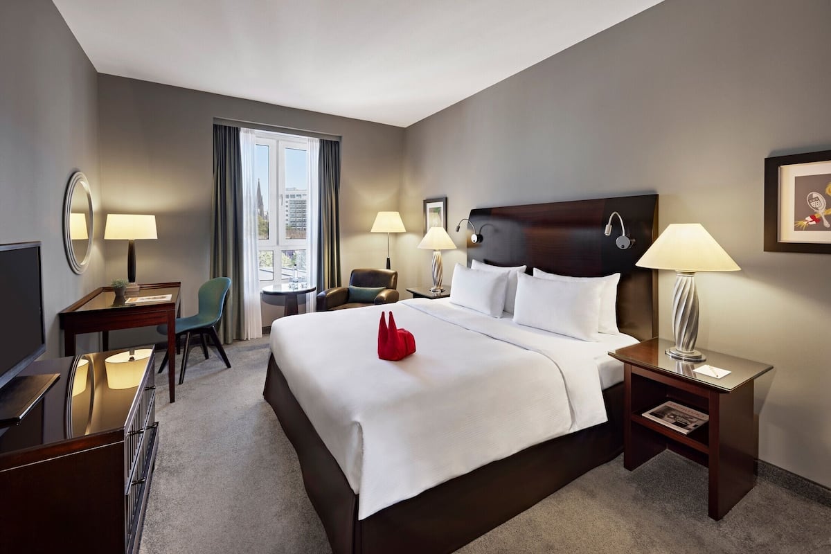 Ein Hotelzimmer mit einem großen Bett, Nachttischlampen, einem Schreibtisch mit Stuhl, einem Fernseher und einem Fenster mit Vorhängen.