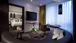 A modern hotel room with a curved sofa, a small round table with a vase of flowers, a flat-screen TV mounted on the wall, and a view of a cathedral through the window.
