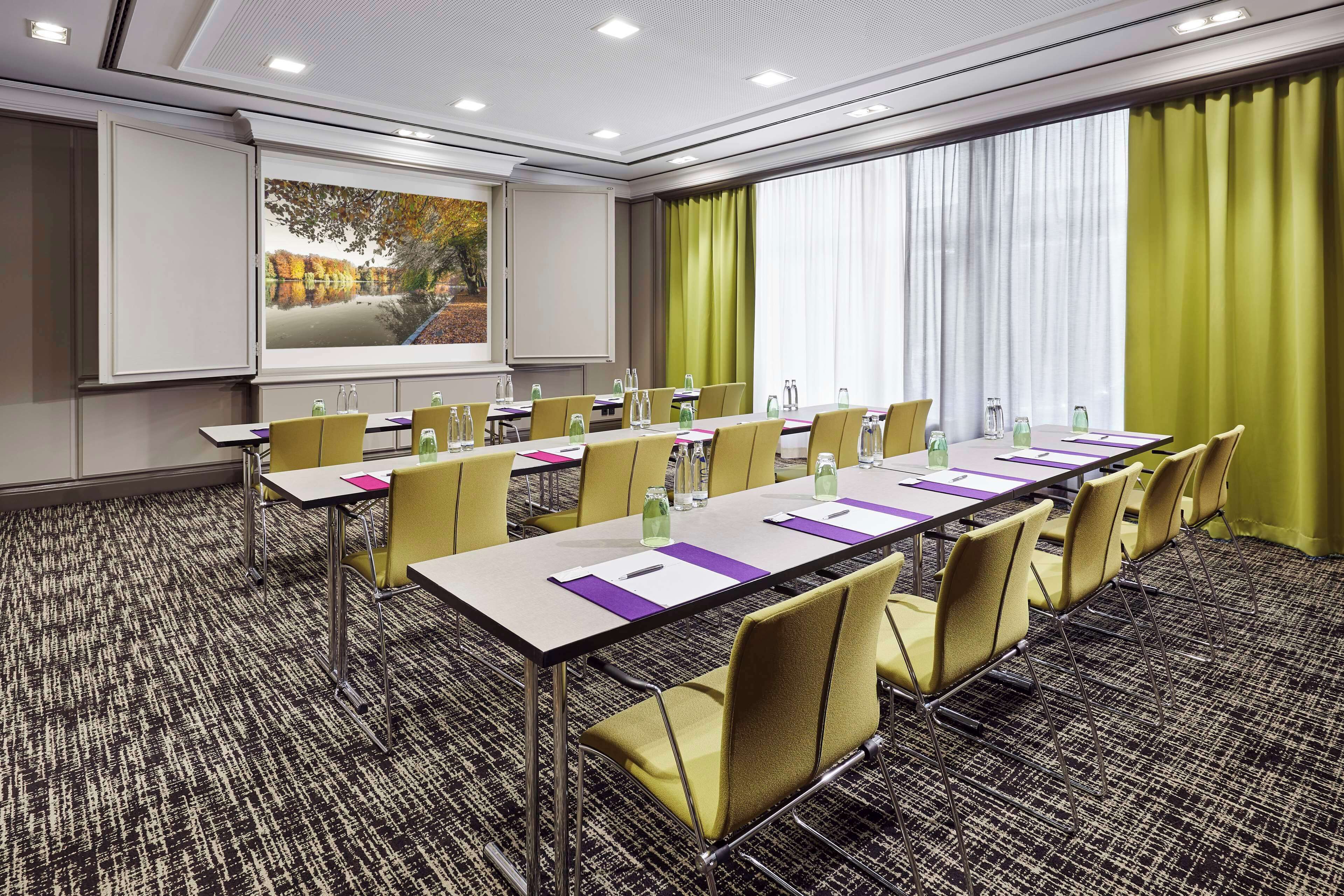 A conference room with a long table, yellow chairs, a projector screen, and a large window with curtains.