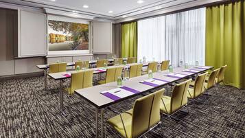 A conference room with a long table, yellow chairs, a projector screen, and a large window with curtains.