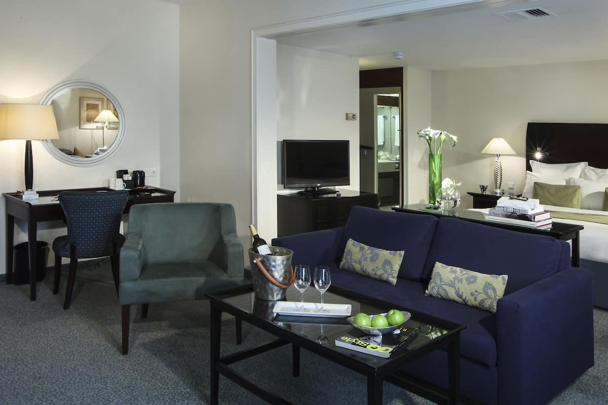 Ein Hotelzimmer mit einer Couch, einem Couchtisch mit einer Zeitschrift, einem Fernseher, einem Bett mit Kissen und einem durch eine geöffnete Tür einsehbaren Badezimmer.