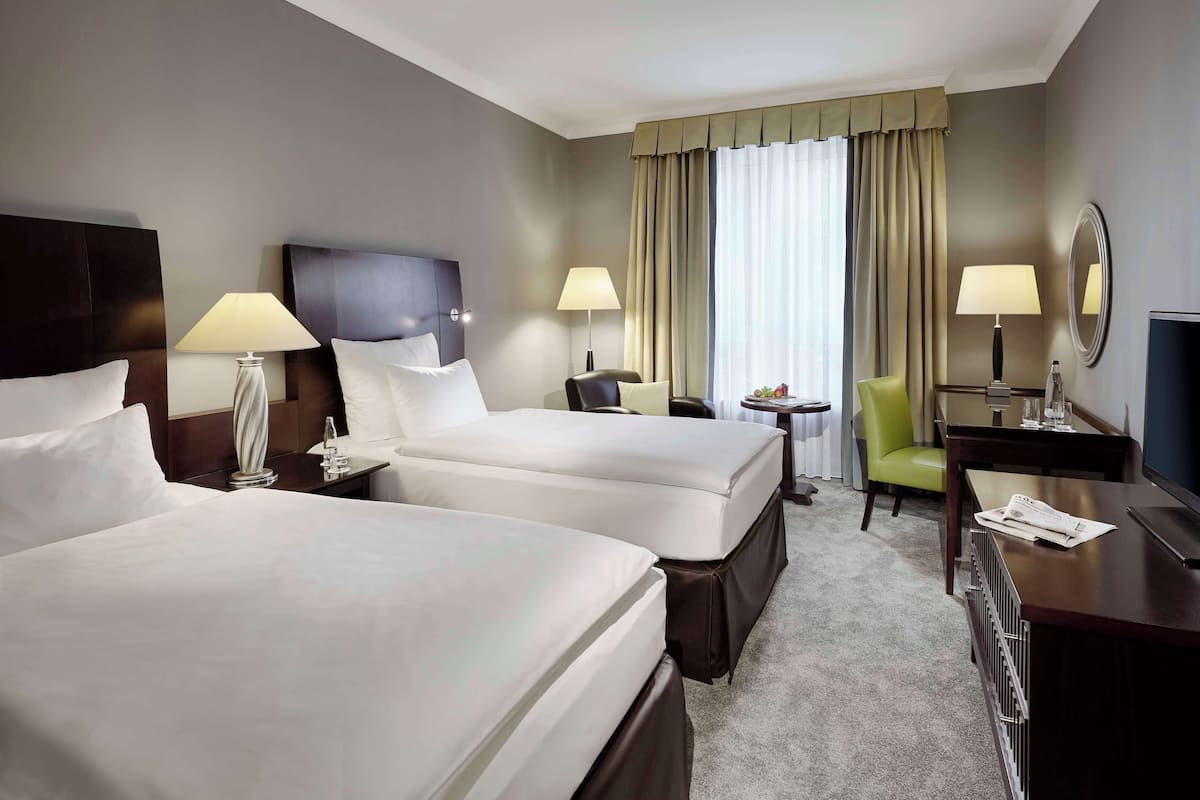 Ein Hotelzimmer mit zwei Betten, einem Schreibtisch, einem Sessel, einem Fernseher und einem Fenster mit Vorhängen.