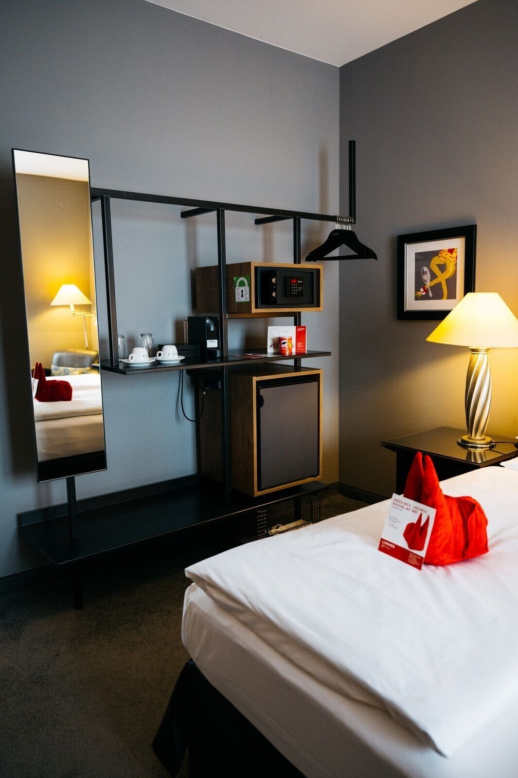 A hotel room with a bed, a desk with a lamp, a mirror, a minibar, and a framed picture on the wall.