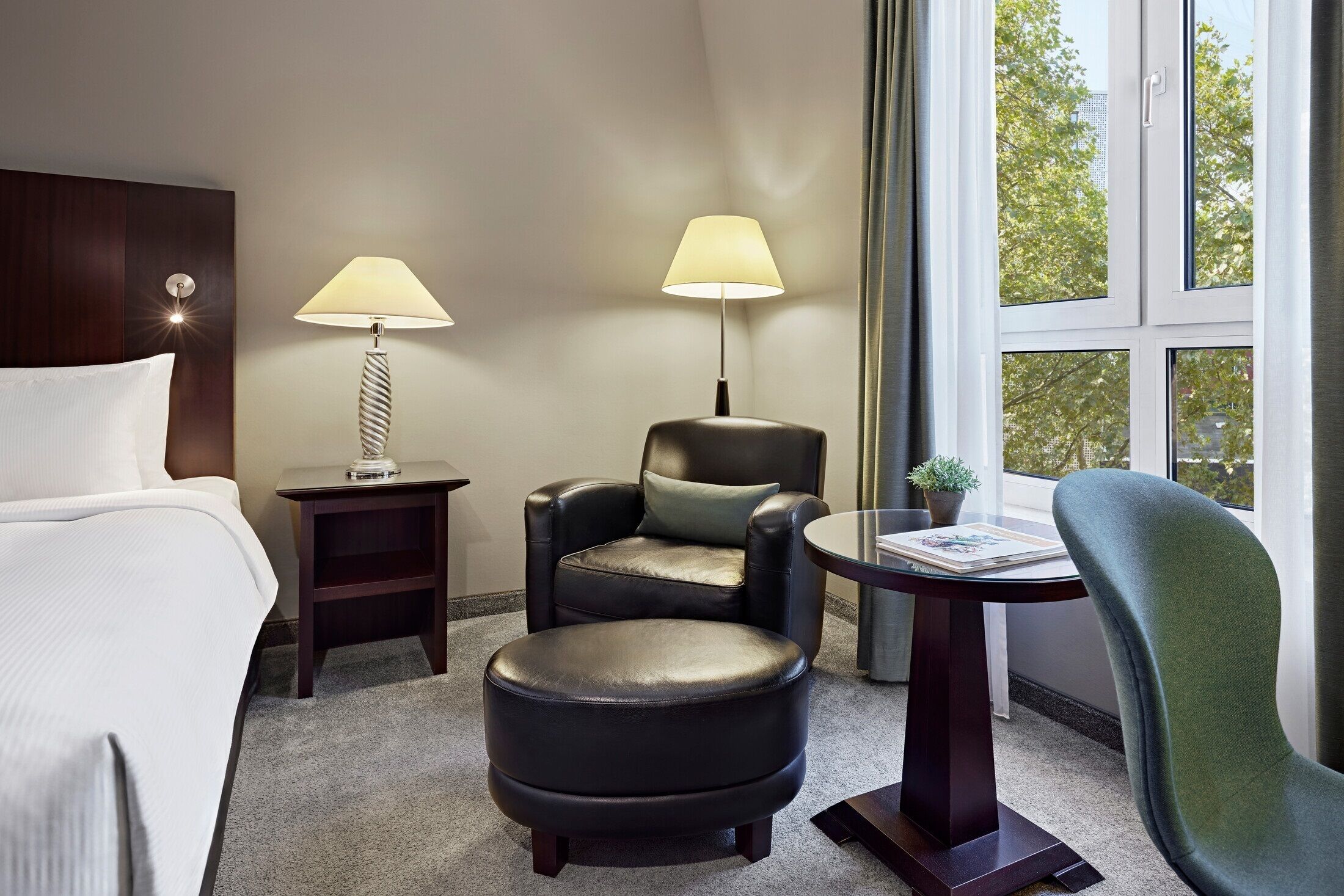 A hotel room with a bed, a desk with a lamp, a chair, an ottoman, and a view of trees through the window.