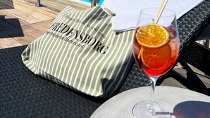 A poolside setting with a glass of red drink, a striped towel, and a cushion with text.