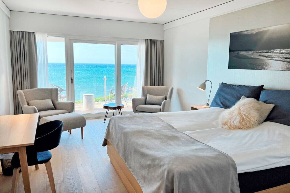 A modern hotel room with a large bed, two armchairs, a small table, and a view of the ocean.