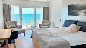 A modern hotel room with a large bed, two armchairs, a small table, and a view of the ocean.