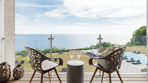 A balcony with two chairs, a small table, and two wine glasses. The view includes a pool and a body of water.
