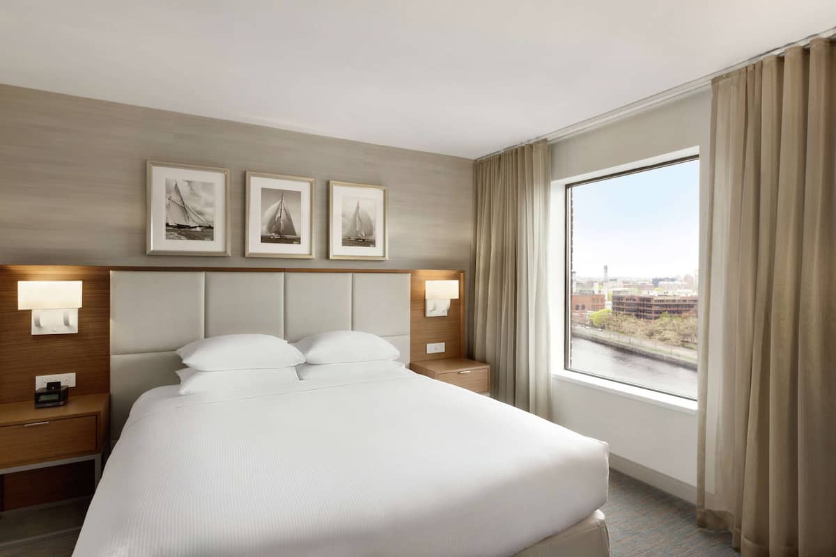 Een hotelkamer met een groot bed, twee nachtkastjes met lampen, een raam met uitzicht op een stadsgezicht en drie ingelijste foto’s aan de muur.