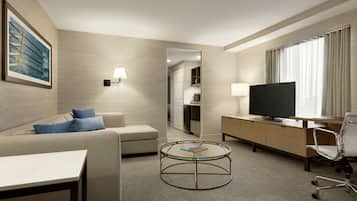 A modern hotel room with a sofa, a glass-top coffee table, a TV, a desk, and a chair.