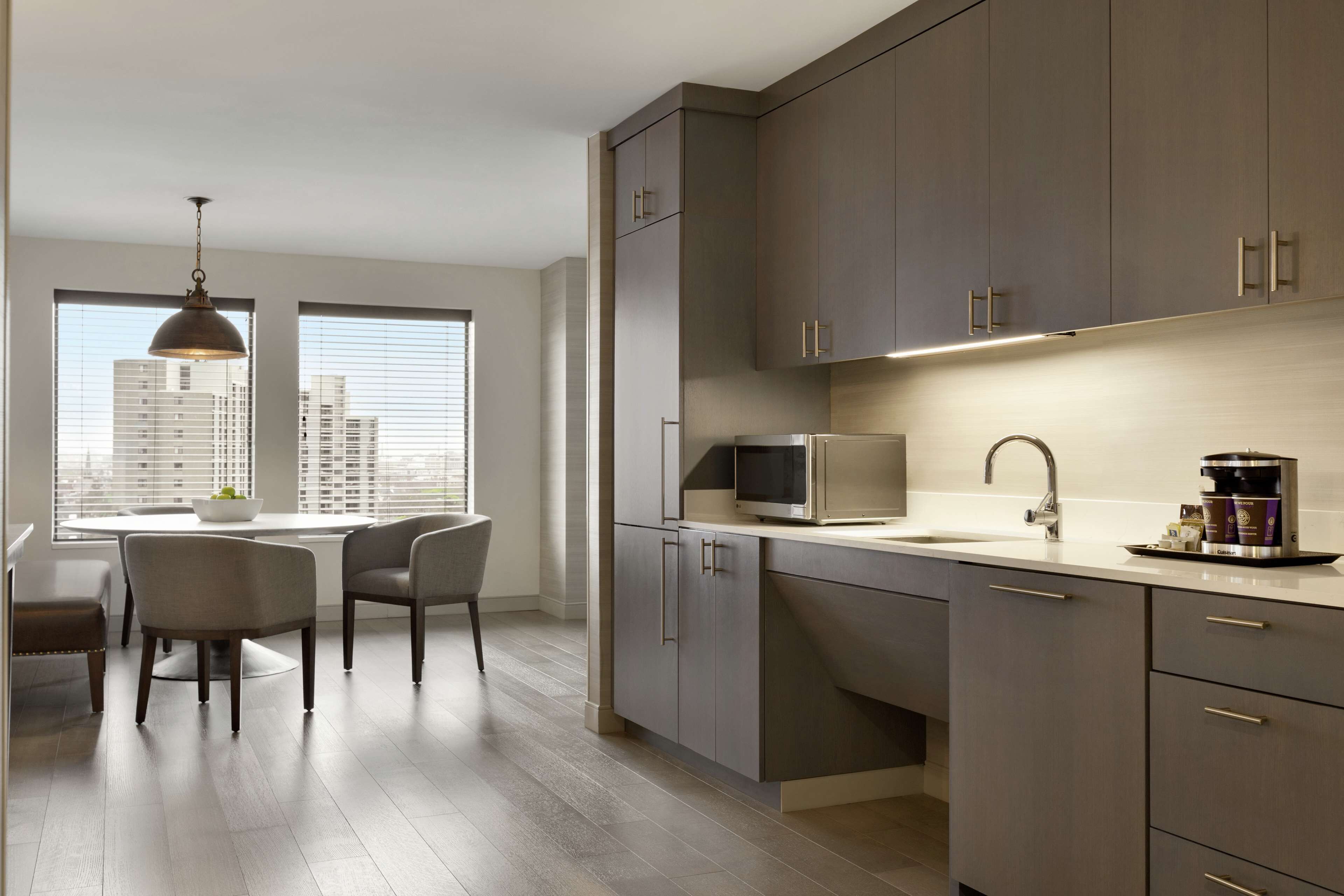A modern kitchen with grey cabinets, a microwave, and a coffee maker on the counter. There is a dining table with chairs and a pendant light above it.