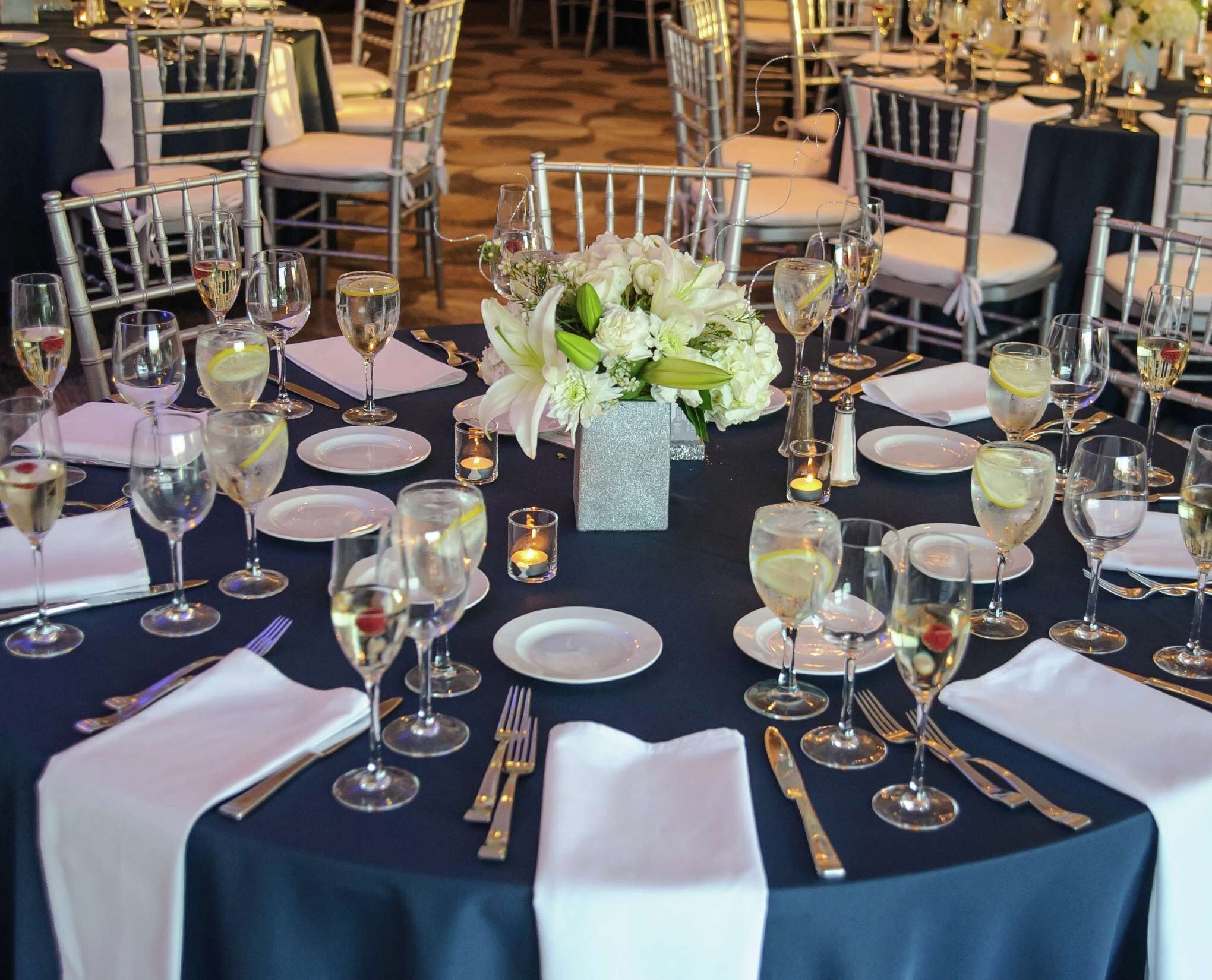 Mesa de comedor formal dispuesta con platos blancos, copas de vino y cubiertos, rodeada de sillas con cojines blancos.