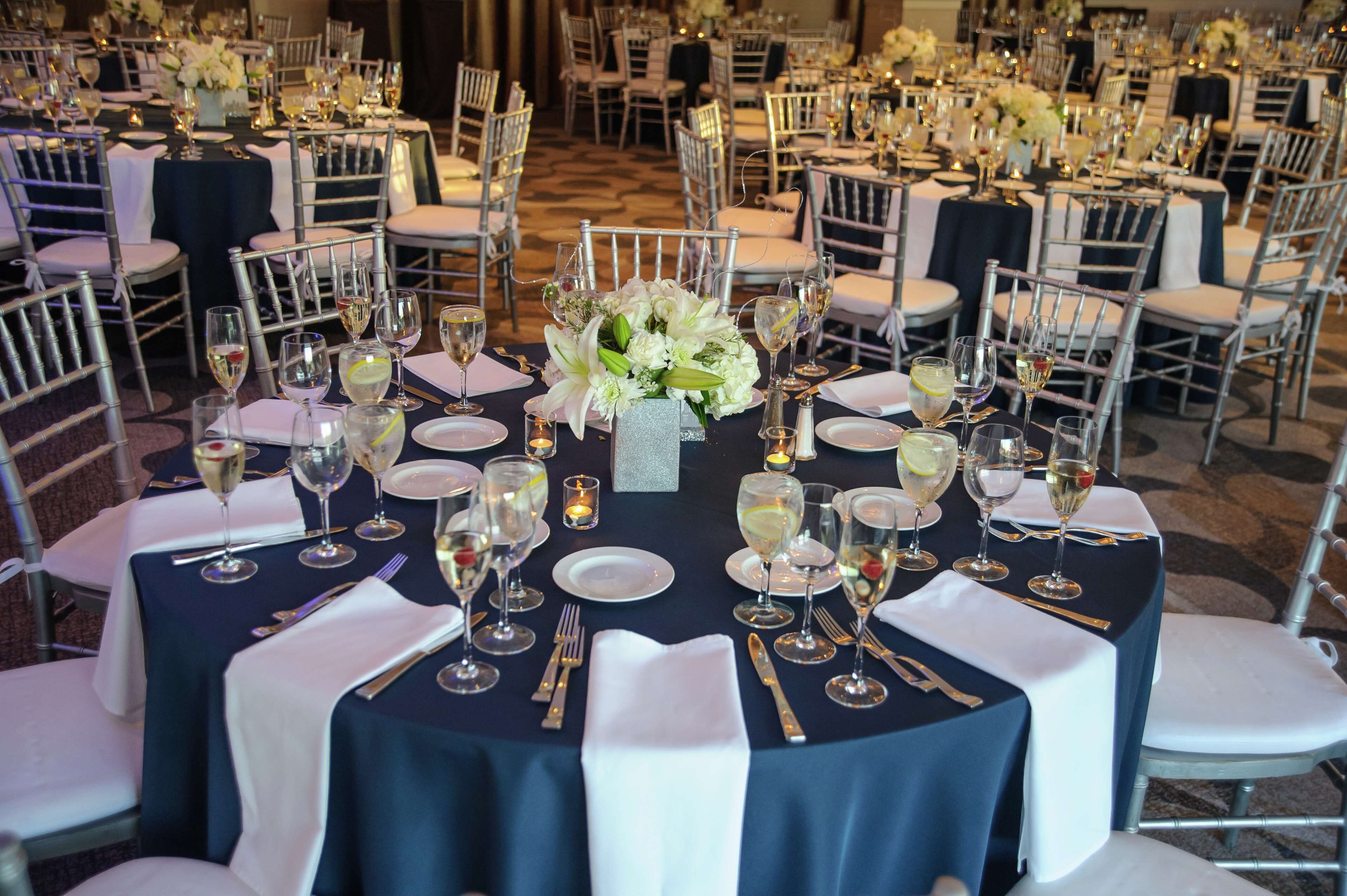 A banquet hall with round tables set for a formal event, featuring white chairs, dark tablecloths, and floral centerpieces.
