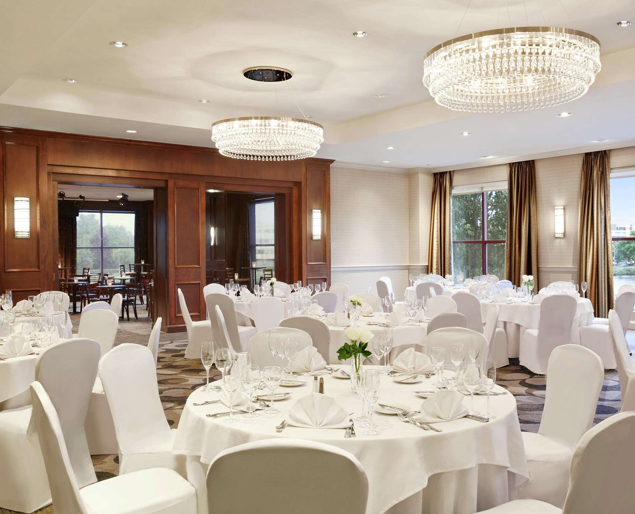 A banquet hall with round tables set for dining, large chandeliers, and a view of the outdoors through large windows.