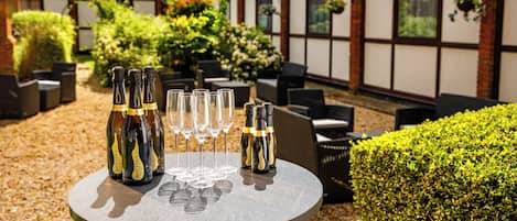 A round outdoor table with bottles and glasses, surrounded by outdoor seating and greenery.
