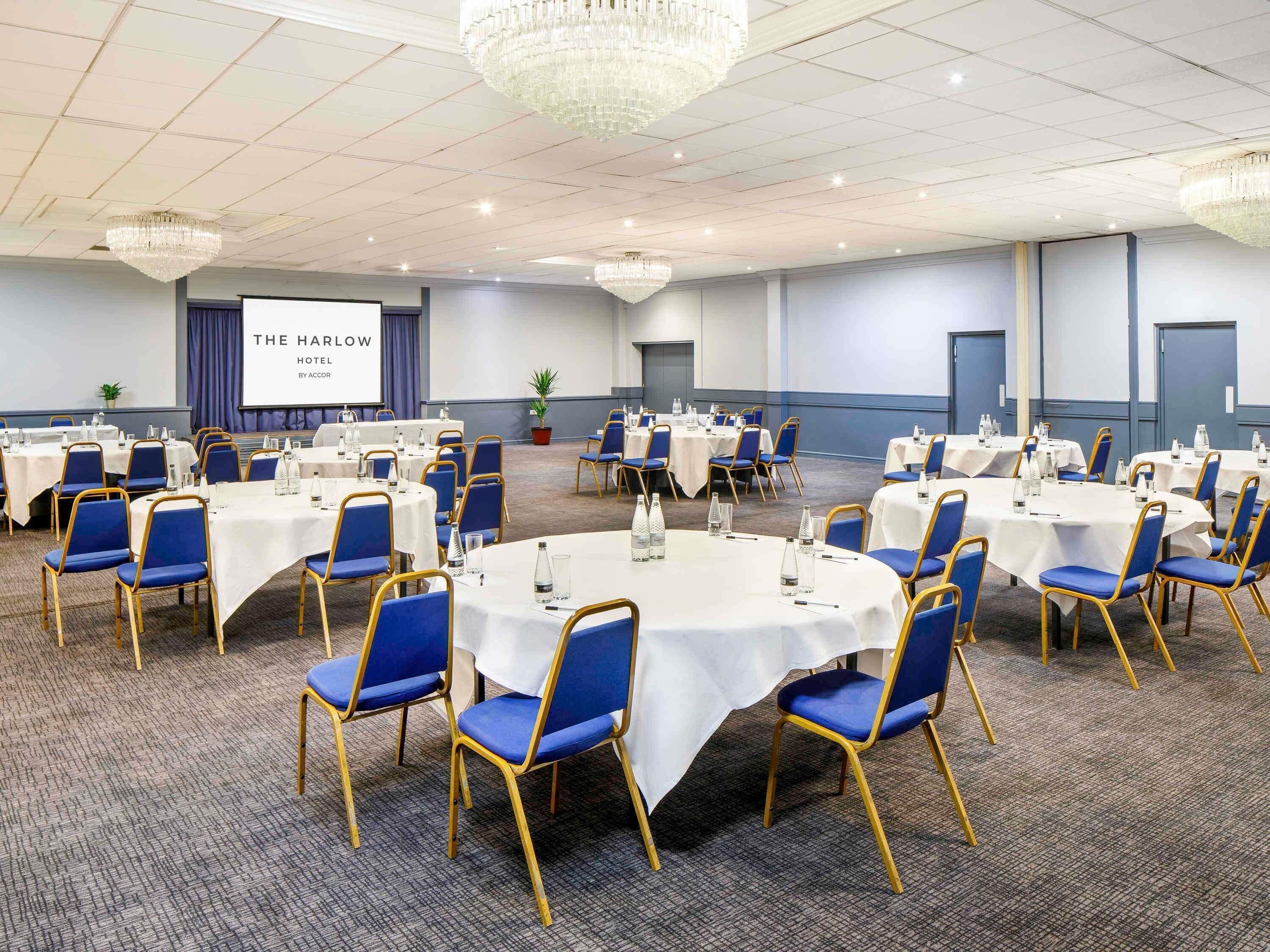 A conference room with round tables, blue chairs, and a projector screen displaying 'THE HARLOW HOTEL BY ACCOR'.