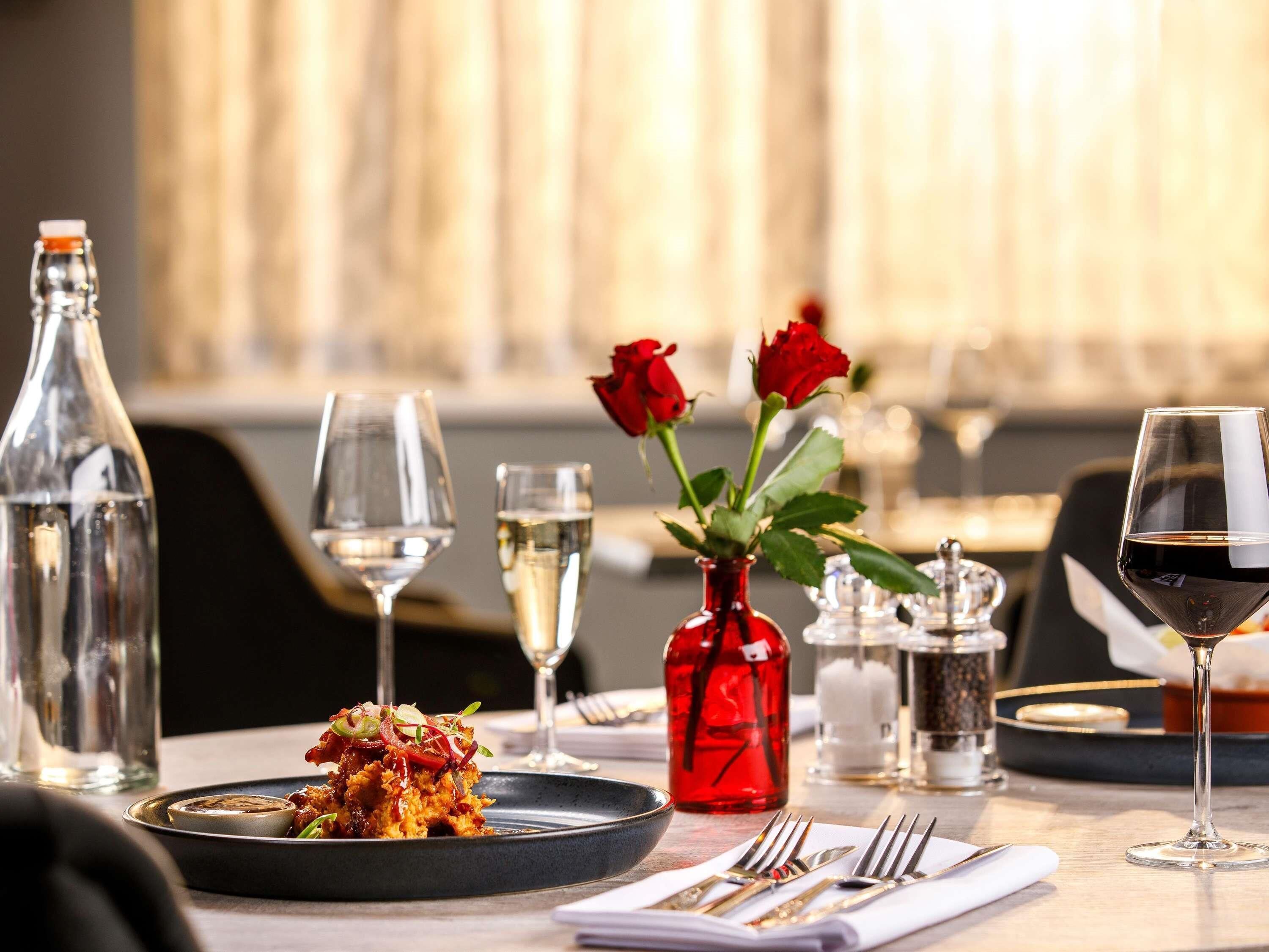 A dining table set with a plate of food, wine glasses, a red vase with roses, and a bottle.