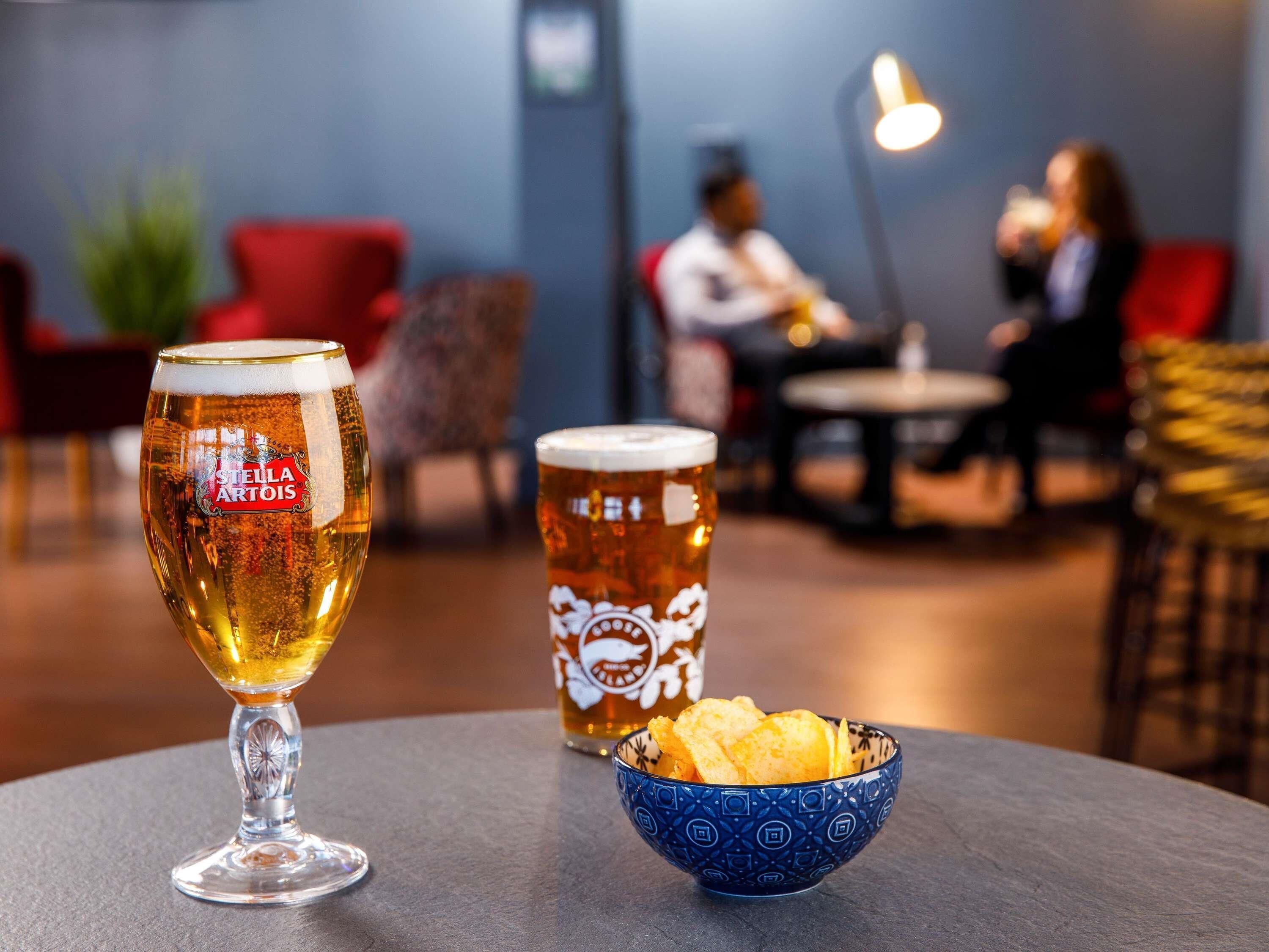 A bar setting with a glass of beer, a mug, and a bowl of chips on a table.