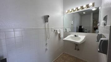 Um banheiro com paredes azulejadas brancas, pia, espelho com iluminação, secador de cabelo e secador de mãos.