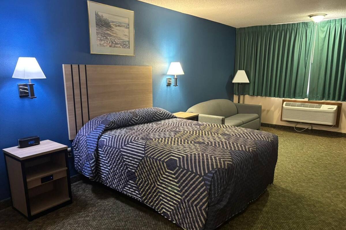Una habitación de hotel con una cama, dos lámparas de noche, una silla, un escritorio, un reloj, un cuadro en la pared y un aire acondicionado.
