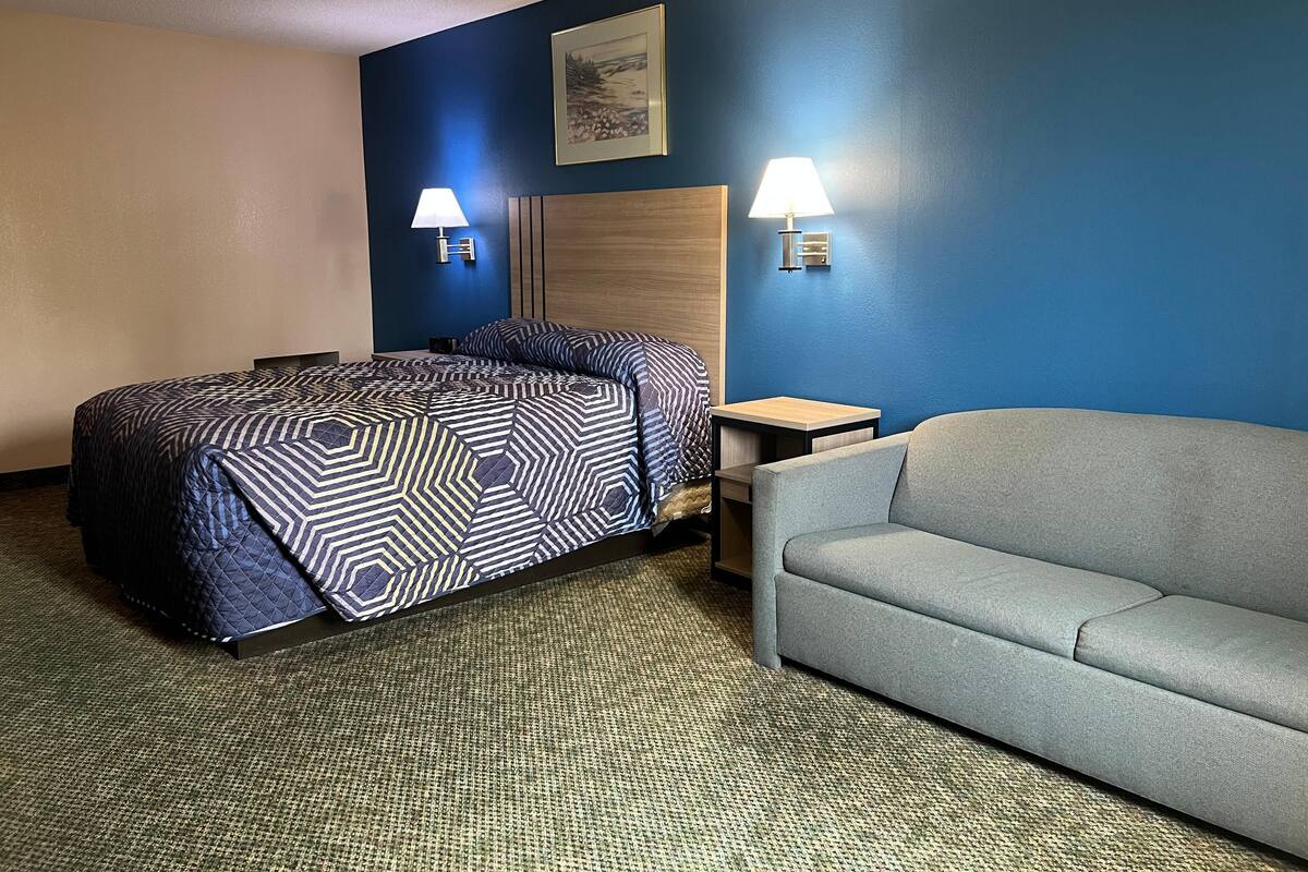 Habitación de hotel con cama, sofá gris, dos lámparas fijadas a la pared y un cuadro enmarcado.