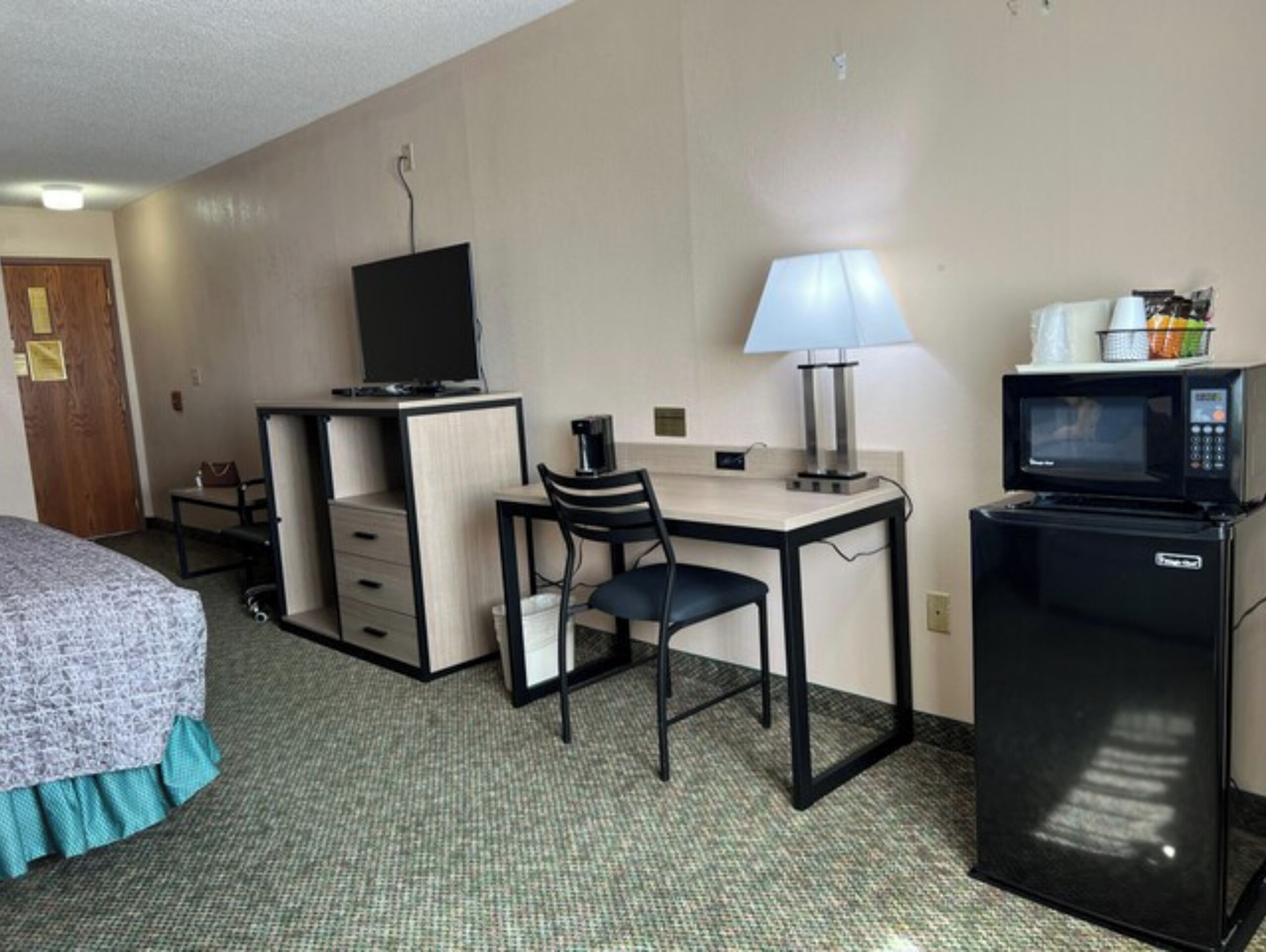 Habitación de hotel con cama, escritorio, silla, televisión, lámpara y refrigerador.