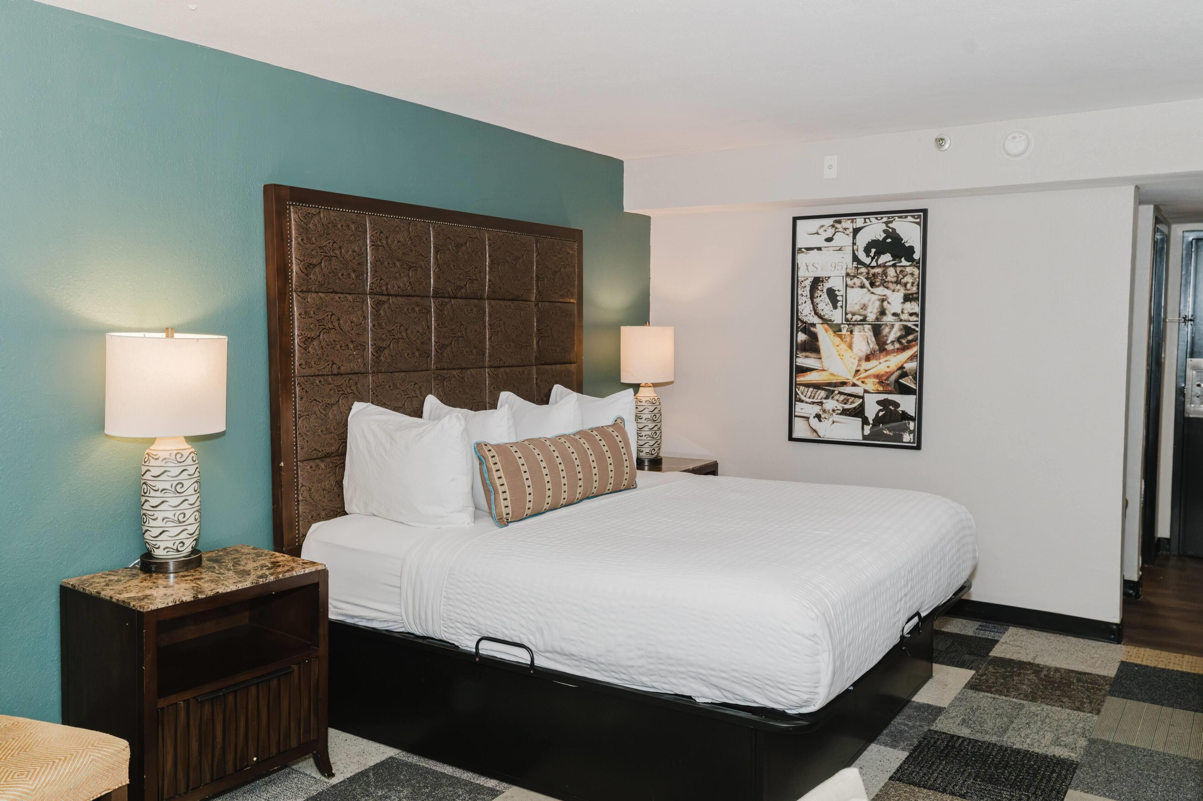 A hotel room with a bed, two bedside lamps, a nightstand, a wall-mounted artwork, and a door leading to another room.