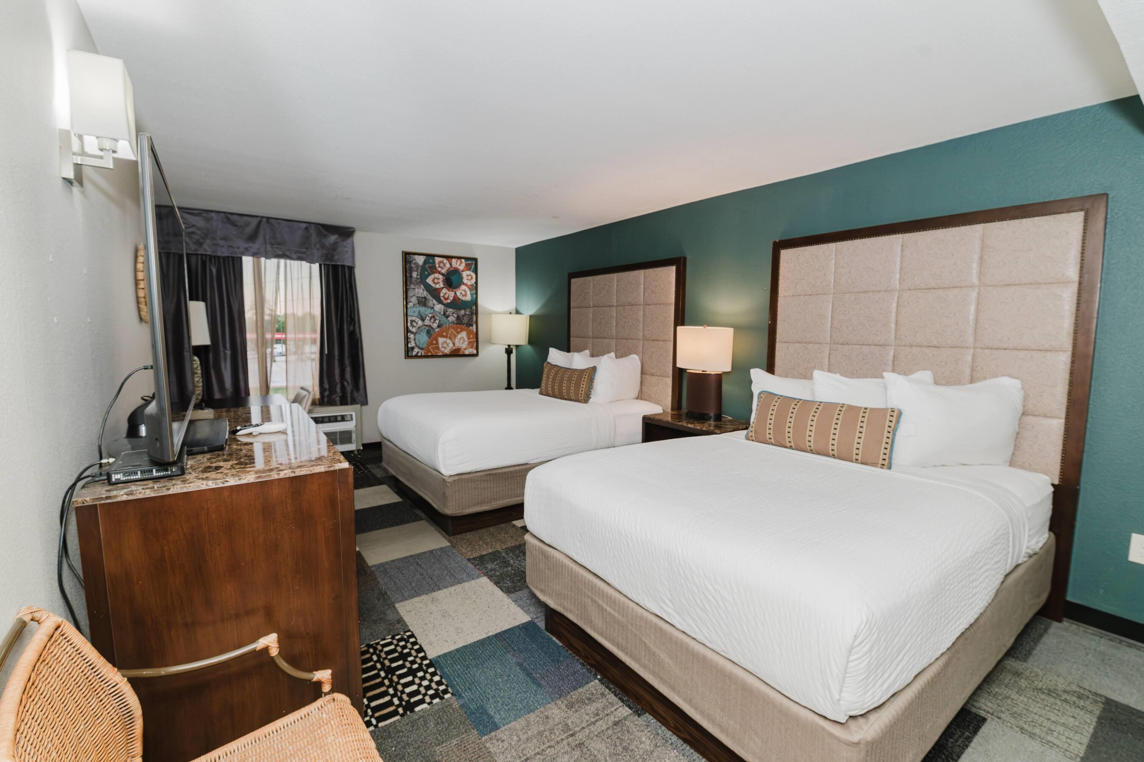 A hotel room with two beds, a desk, a chair, a TV, and a window with curtains.