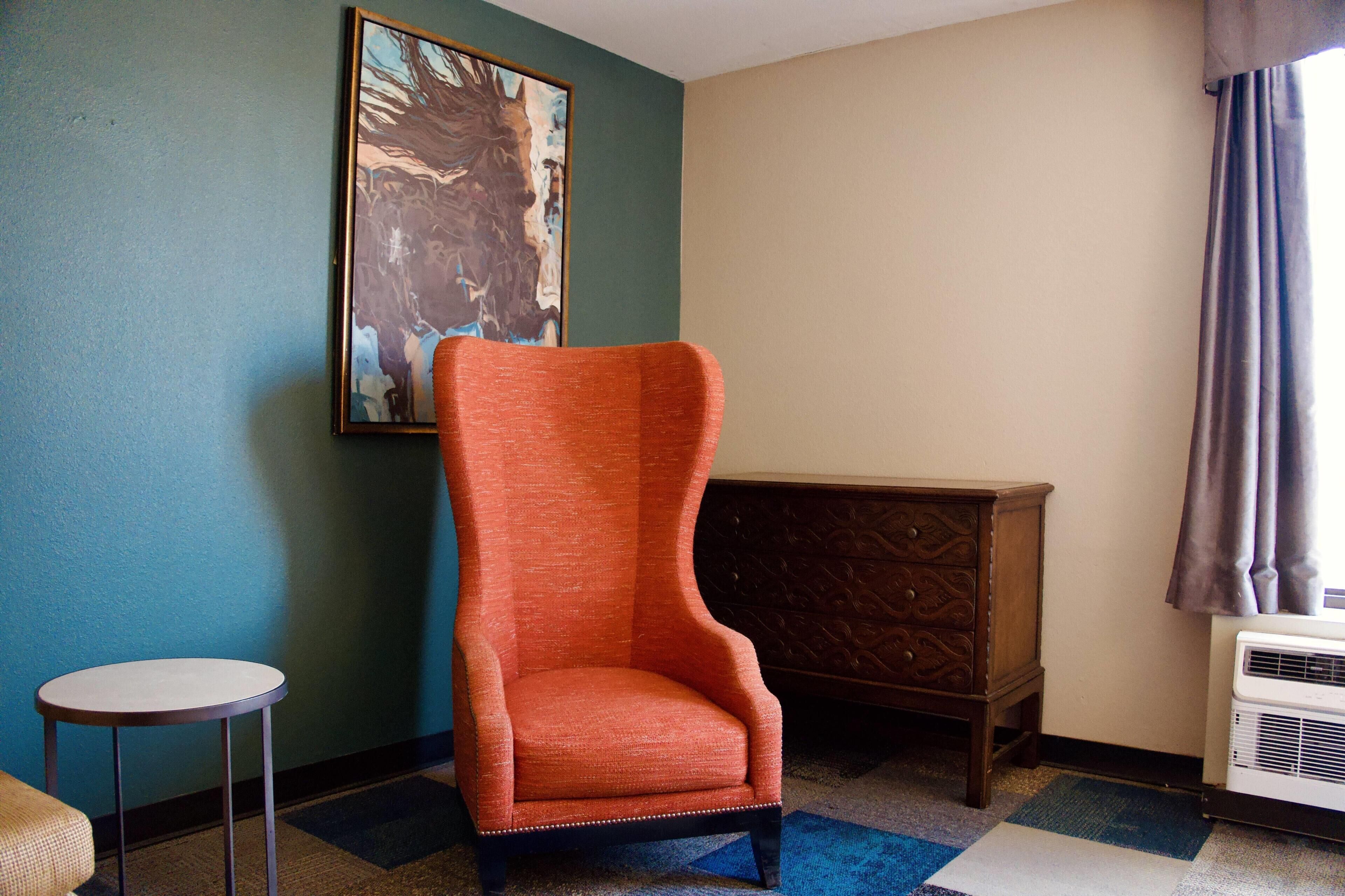 A room with a single orange armchair, a small round table, a wooden chest of drawers, and a large painting on the wall.