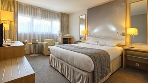 A hotel room with a large bed, a desk with a chair, a small table, and a mirror. - Hôtel Inn Paris CDG Airport (Roissy-en-France)
