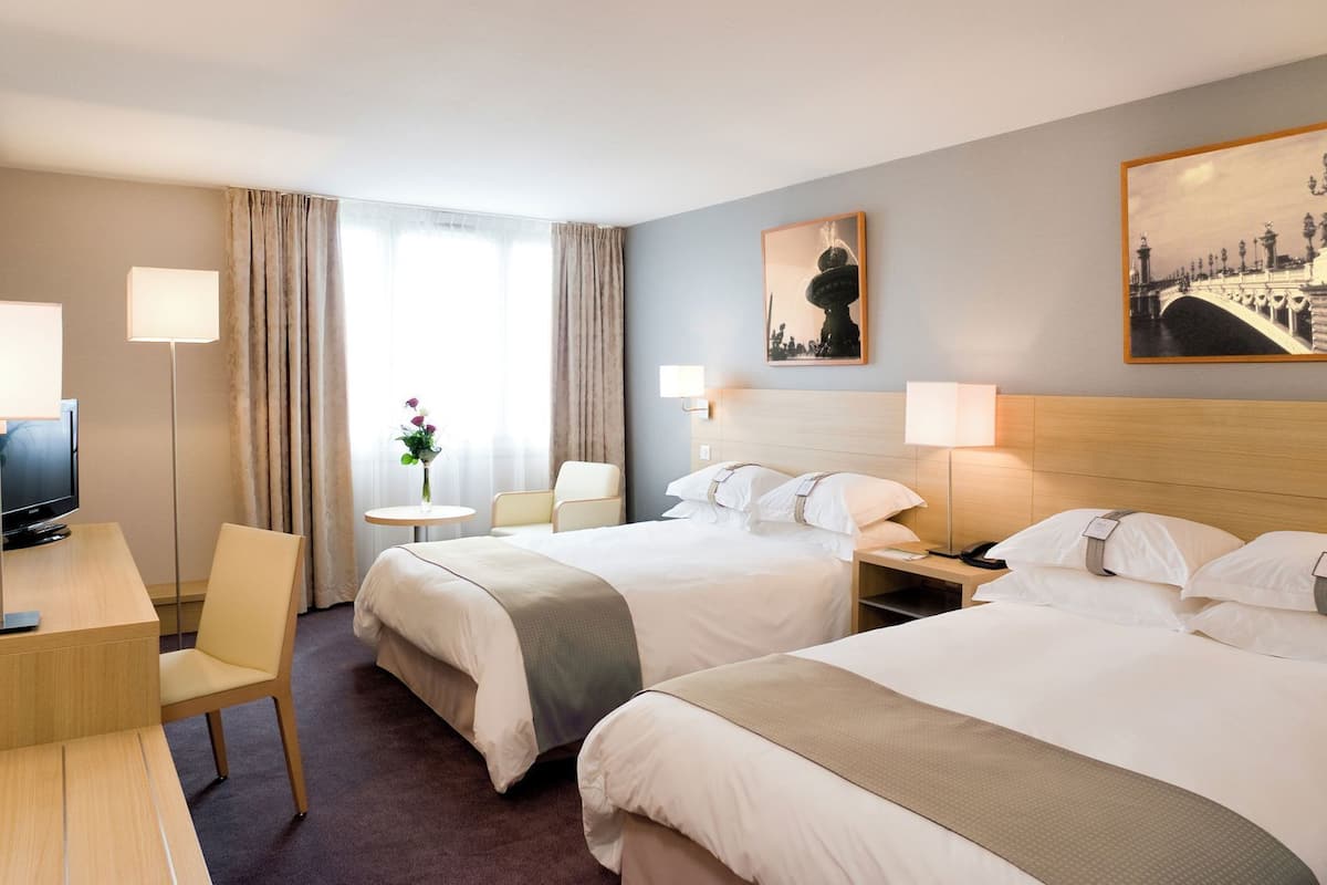 Ein Hotelzimmer mit zwei Betten, einem Schreibtisch mit Fernseher, einem Sessel, einem kleinen Tisch mit Blumenvase und zwei Wandleuchten.