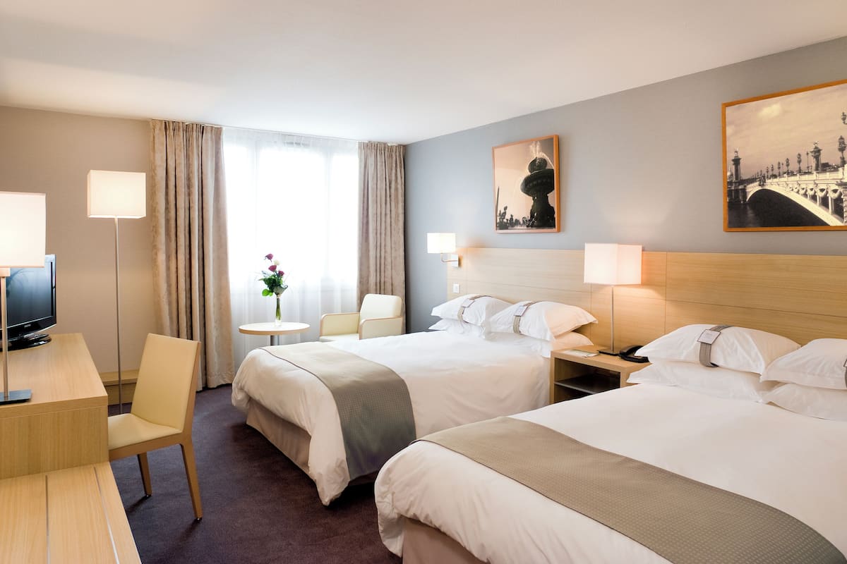 Ein Hotelzimmer mit zwei Betten, einem Schreibtisch, einem Sessel, einem Fernseher und einem Fenster mit Vorhängen.