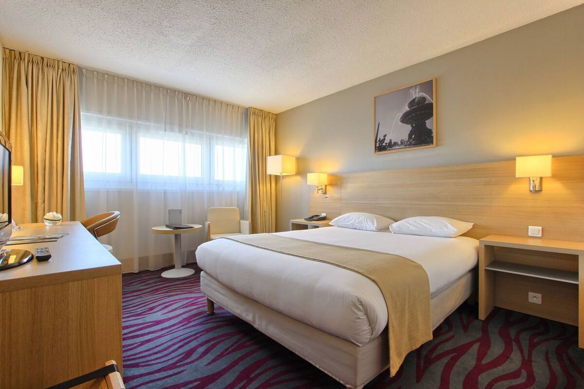 Ein Hotelzimmer mit einem Bett, Nachttischen, einem Schreibtisch mit Stuhl, einem kleinen Tisch mit einem Laptop und einem Bild an der Wand.