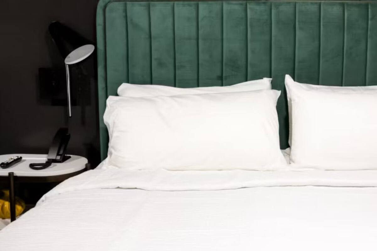 Una cama con ropa de cama blanca, un cabecero tapizado en verde y una mesita de noche con una lámpara.