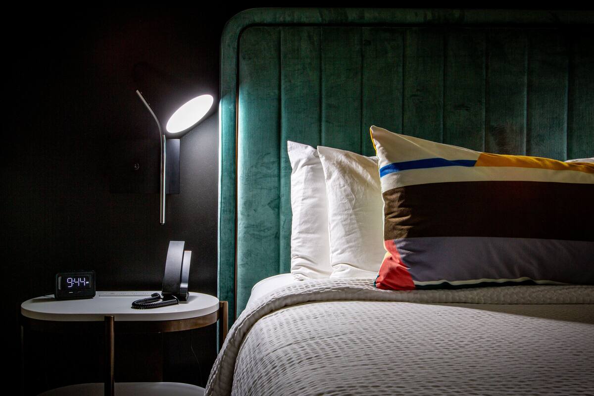 Una cama bien hecha con un cabecero verde, una mesita de noche con lámpara y un reloj digital.