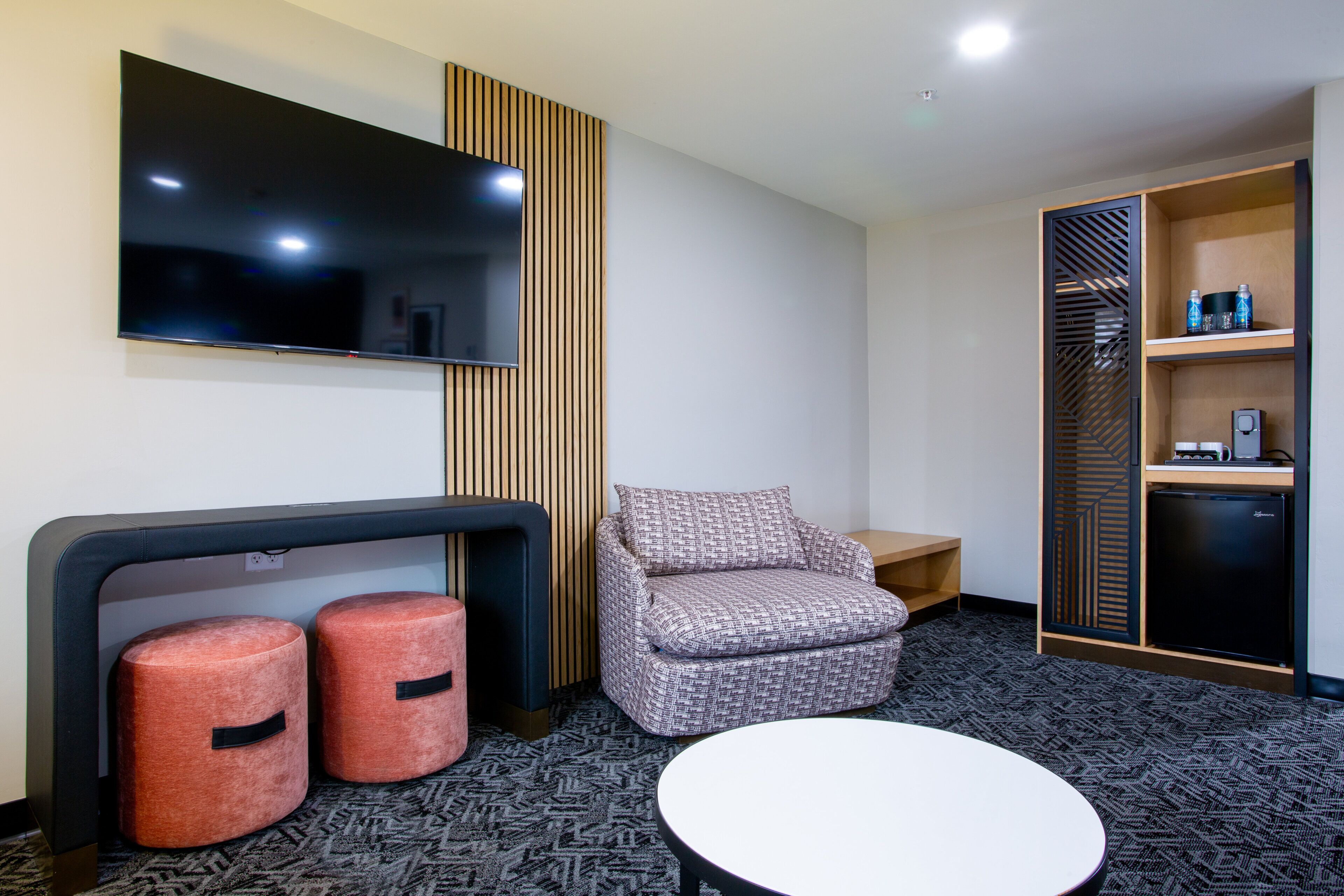 A modern hotel room with a flat-screen TV mounted on the wall, a patterned sofa, two ottomans, a small table, and a built-in closet with shelves.