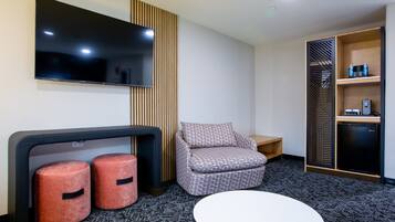 A modern hotel room with a flat-screen TV mounted on the wall, a patterned sofa, two ottomans, a small table, and a built-in closet with shelves.