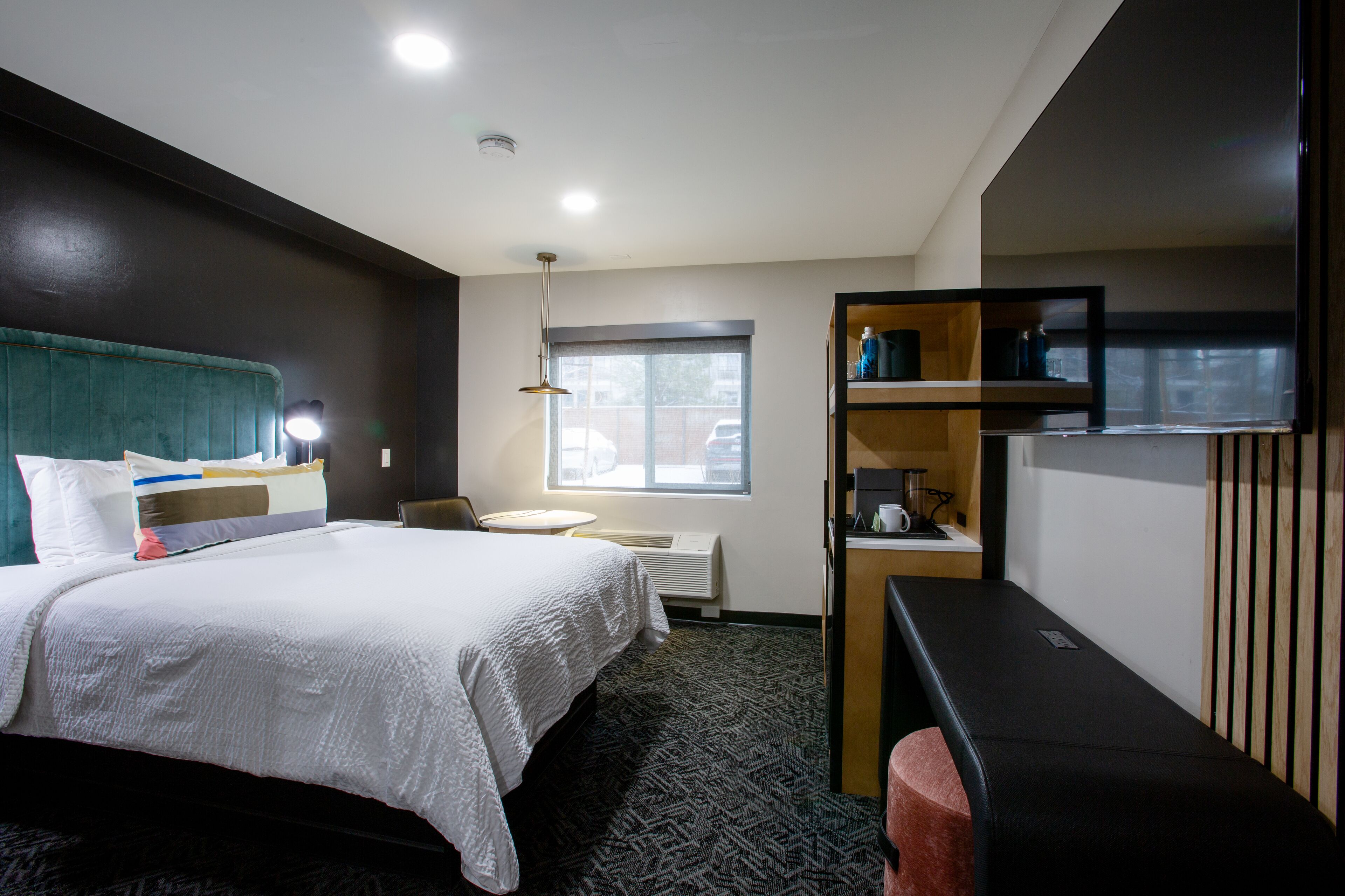 A hotel room with a bed, a desk, a chair, a window, and a shelf with items.