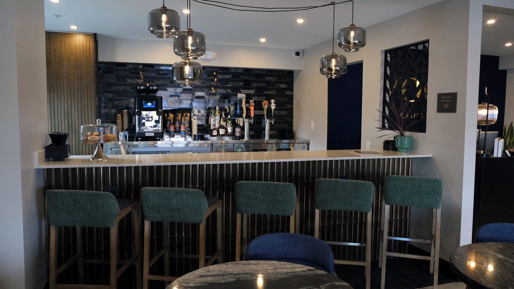 Zona de bar con diseño moderno, que incluye una barra con taburetes, una variedad de botellas y una pantalla digital.