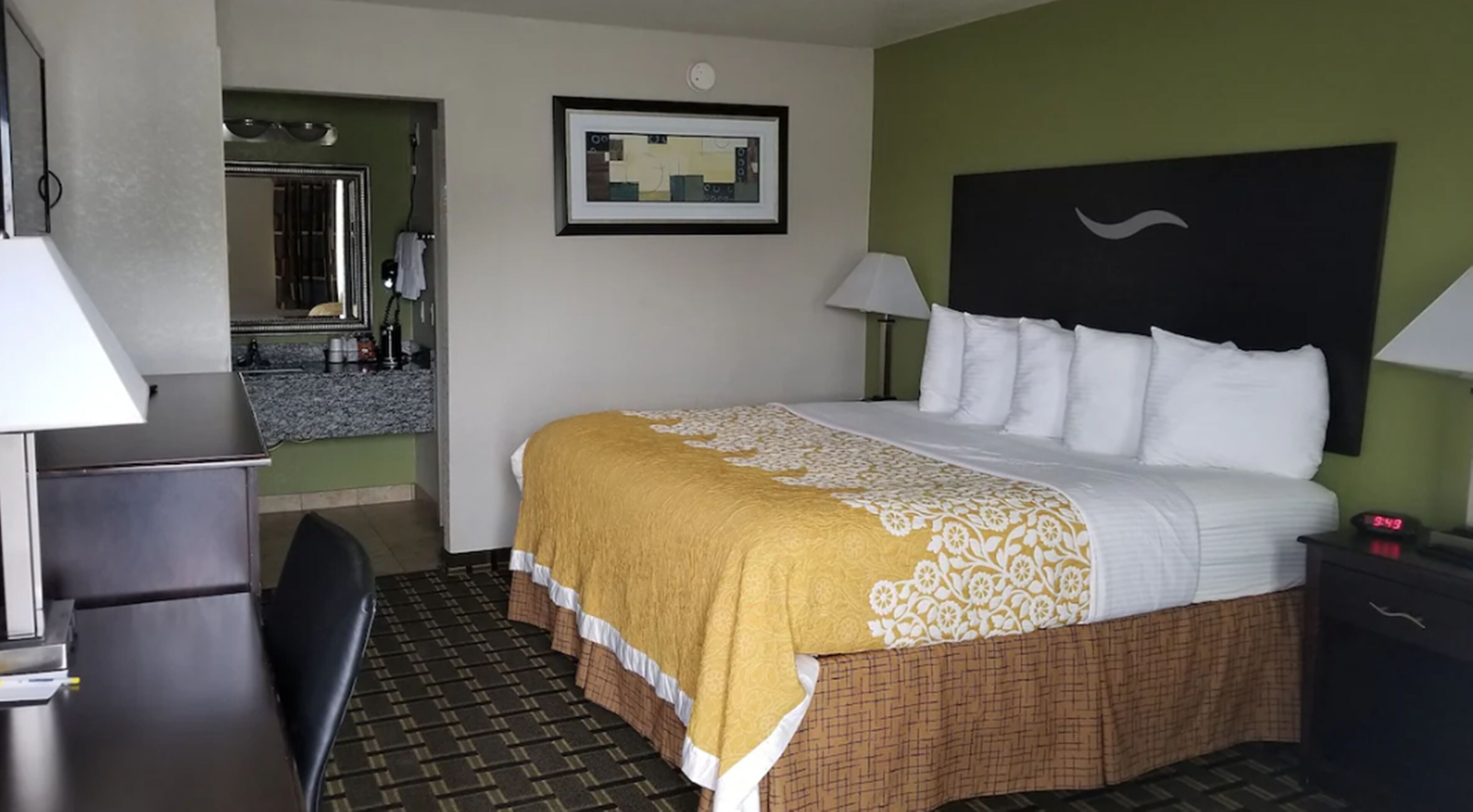 A hotel room with a bed, a desk, a chair, a nightstand, a lamp, a clock, a framed picture, and a bathroom visible through an open door.