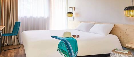 A hotel room with a large bed, a desk, a chair, a lamp, and a woven ottoman.
