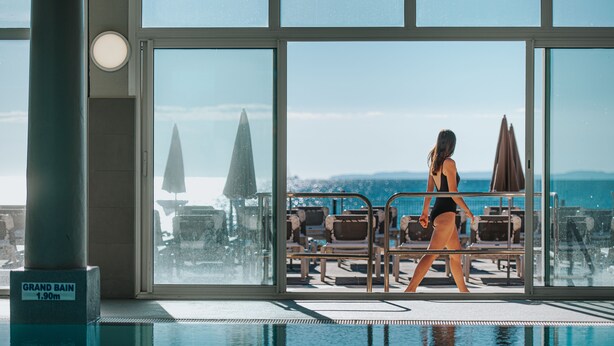 Un espace aménagé au bord de la piscine, avec des chaises longues et des parasols, une grande fenêtre offrant une vue sur la ville, et un panneau indiquant la profondeur de la piscine.